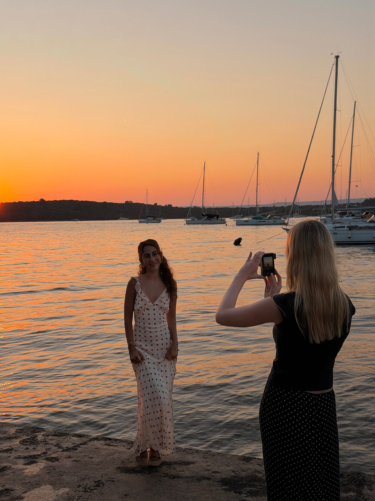 A woman stands on a rocky shoreline at sunset, while another woman takes a photo. Sailboats are anchored on the water behind them. 