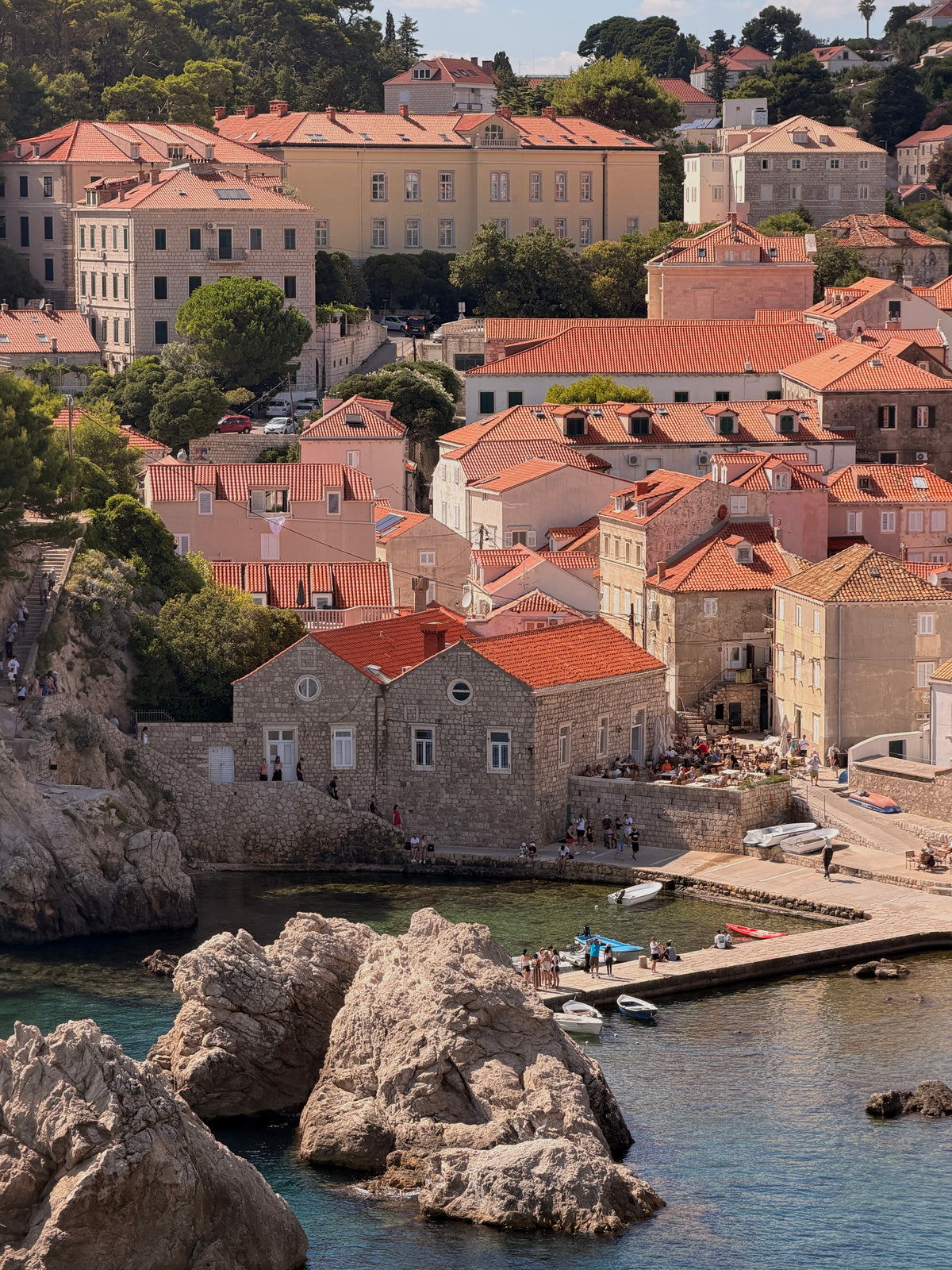 Aerial view of Dubrovnik's Old Town in Croatia, showing a dense cluster of historic stone buildings with distinct orange terracotta roofs, completely surrounded by a massive medieval stone wall and bordered by the deep blue Adriatic Sea.