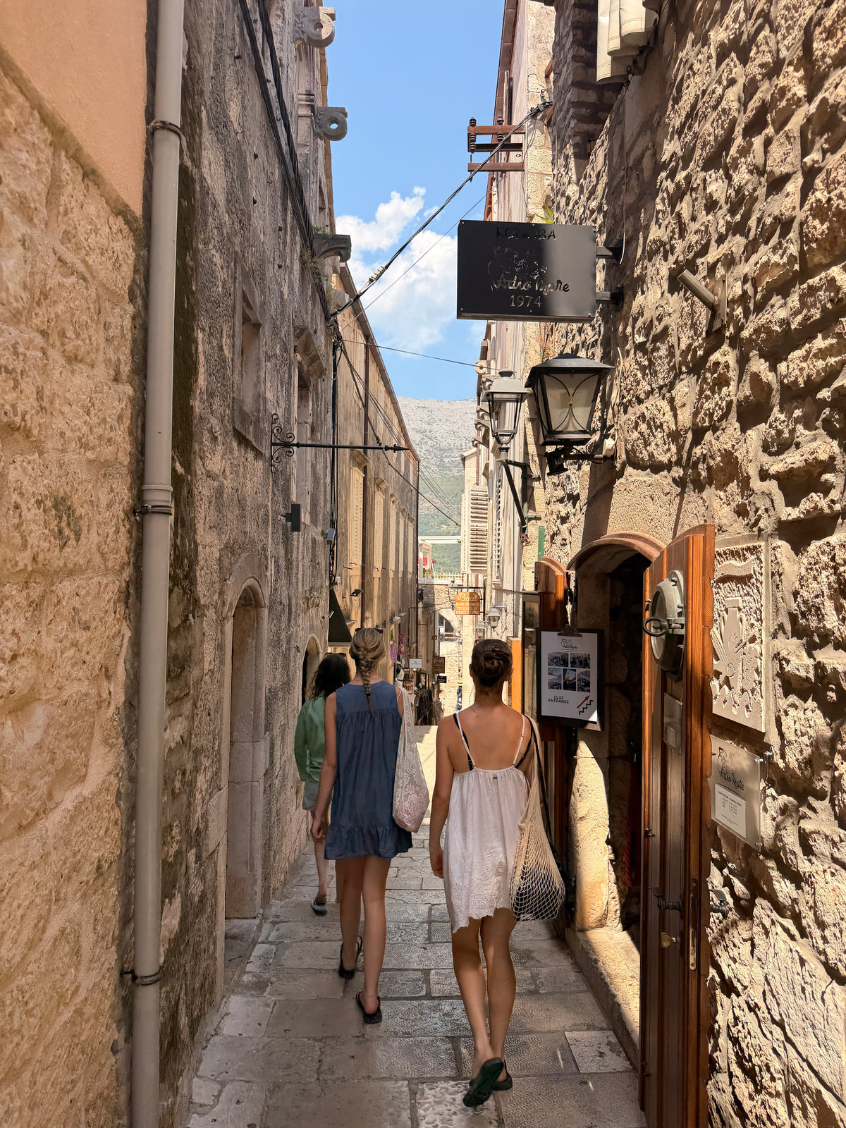 Two women walking down a narrow, sunlit stone alleyway between historic buildings, viewed from behind in Korčula Old Town Croatia.
